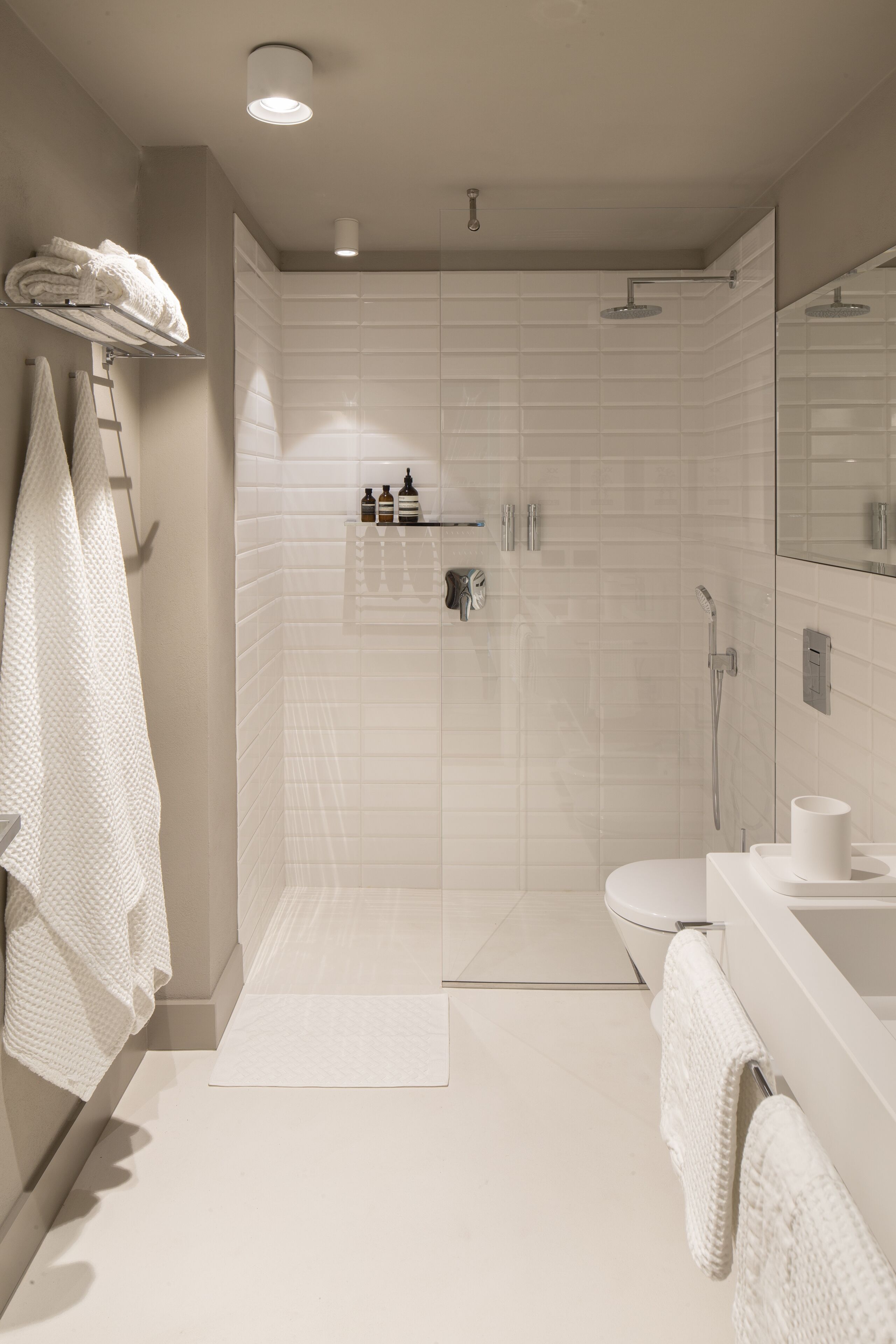 A modern bathroom with a walk-in shower, a toilet, and a sink. There are towels hanging on the wall and a shelf with toiletries.
