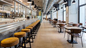 A modern bar area with stools and tables, a bar counter with bottles, and a visible kitchen area. - Edit Lofts  (Turin)