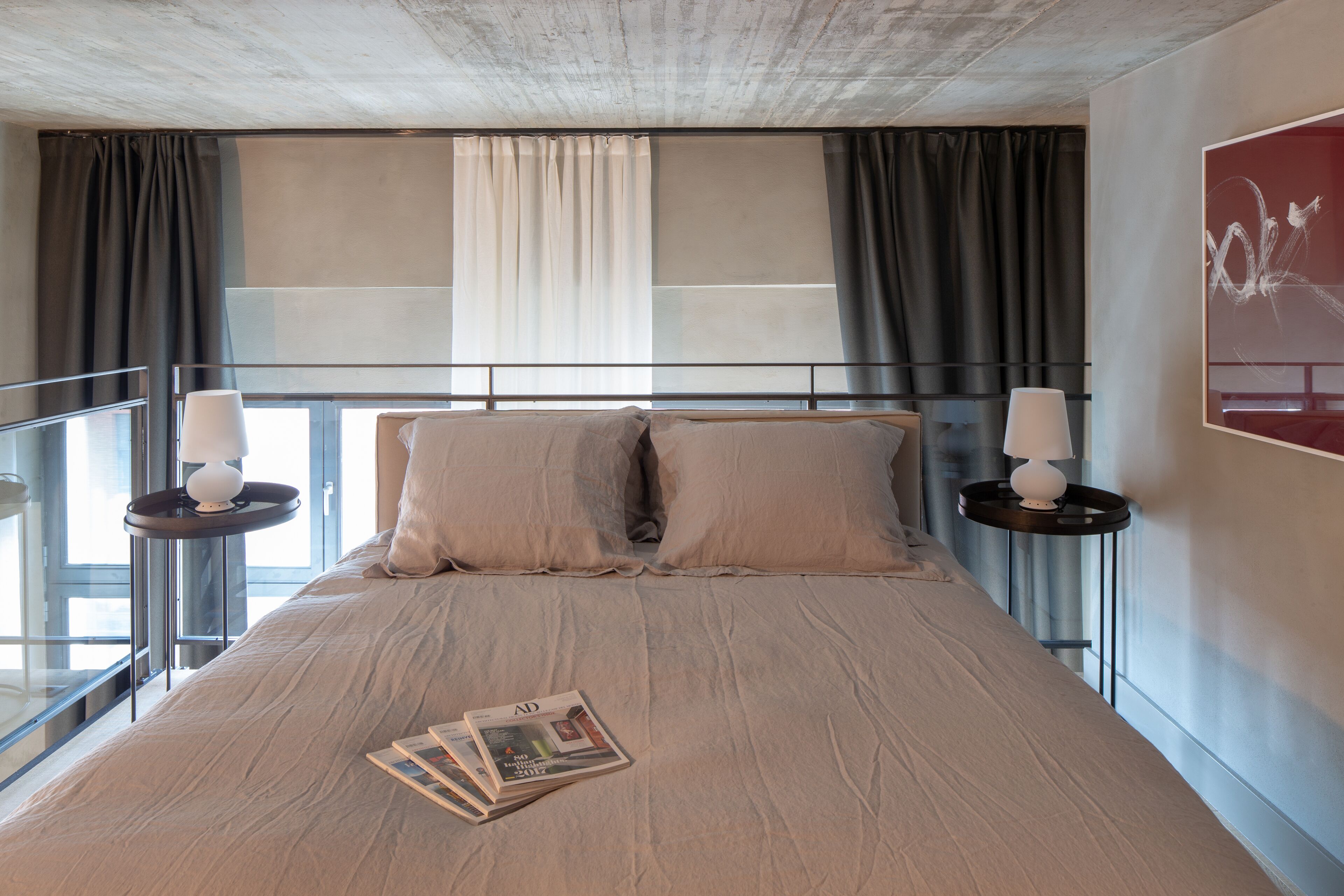 A modern hotel room with a large bed, two bedside tables with lamps, a magazine on the bed, and a view of the city through large windows.