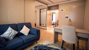 A modern room with a blue sofa, a white chair, a desk, and a glass door leading to a bathroom. - Crowne International Business Hotel (Shiyan)
