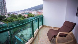 A balcony with a glass railing, a brown chair, and a view of a cityscape with buildings and greenery. - Crowne International Business Hotel (Shiyan)