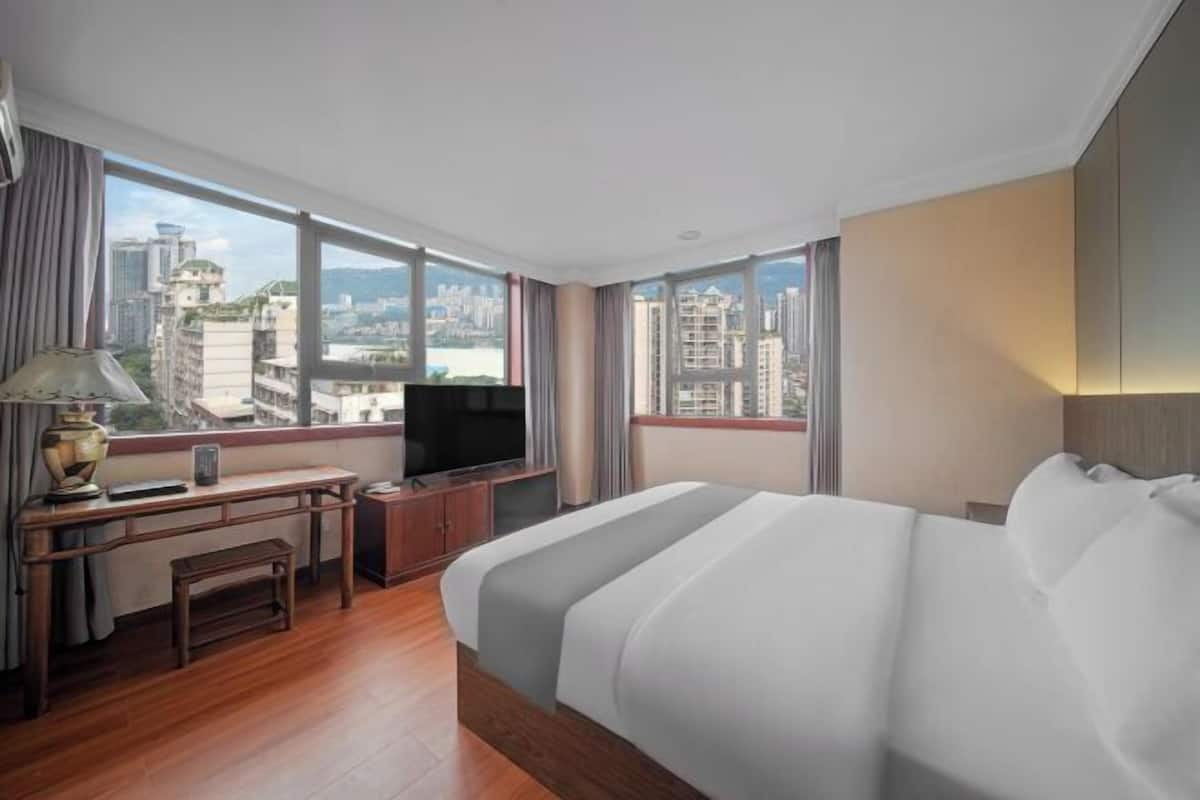 A hotel room with a bed, desk, TV, and large windows offering a city view.