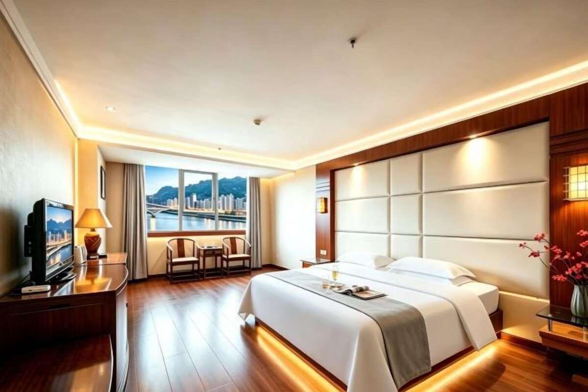 A hotel room with a large bed, a desk with a television, a window with a city view, and a nightstand with a lamp.
