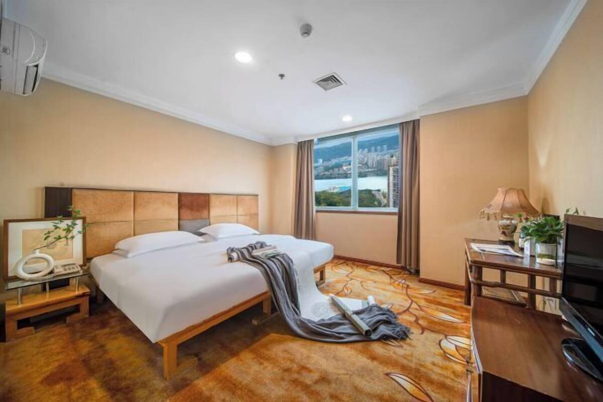 A hotel room with a bed, a desk with a TV, a window with a city view, and a ceiling with recessed lighting.