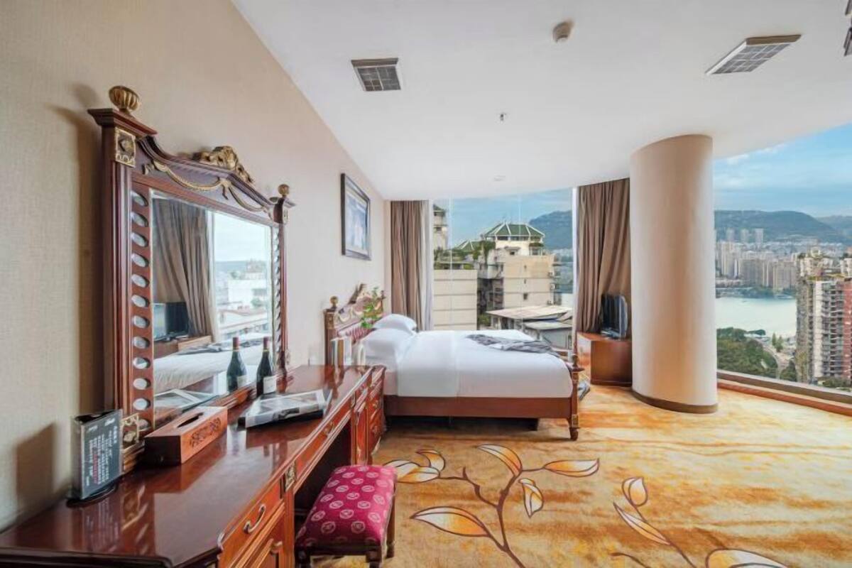 A hotel room with a large bed, a vanity with a mirror, a desk, and a view of a cityscape.