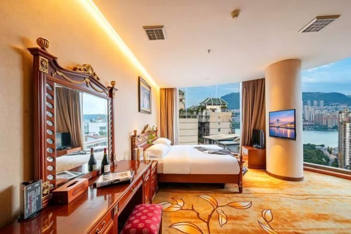 A hotel room with a large bed, a vanity with a mirror, a desk with a chair, a television, and a view of a cityscape and water.