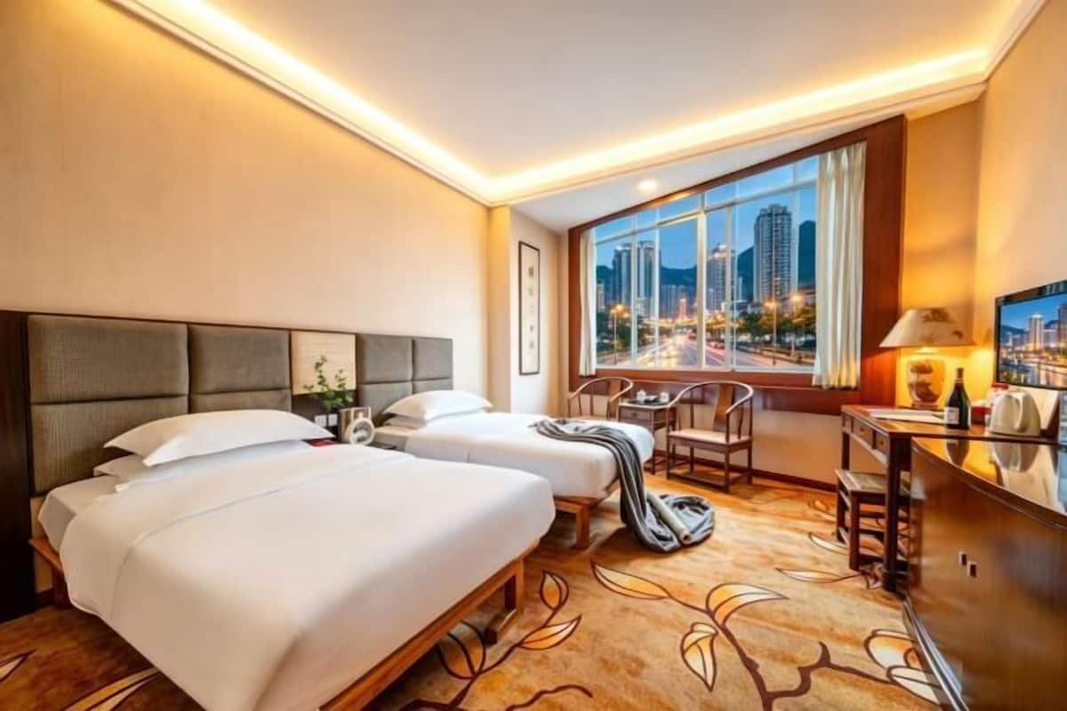 A hotel room with two beds, a desk, a chair, a large window with a city view, and a carpet with a leaf pattern.
