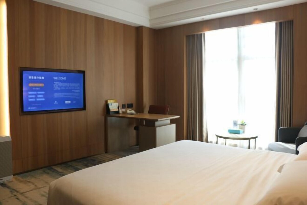 A hotel room with a large bed, a desk, a chair, a television on the wall, and a window with curtains.