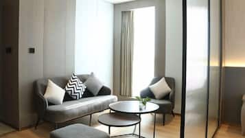 Ein modernes Zimmer mit grauem Sofa, zwei Sesseln, einem kleinen runden Tisch und einem großen Fenster mit Vorhängen.