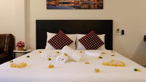 Et hotelværelse med en pænt redt seng, to puder med et prikket mønster, et maleri af en kystby over sengen og en stol med en vase med blomster på sengebordet.