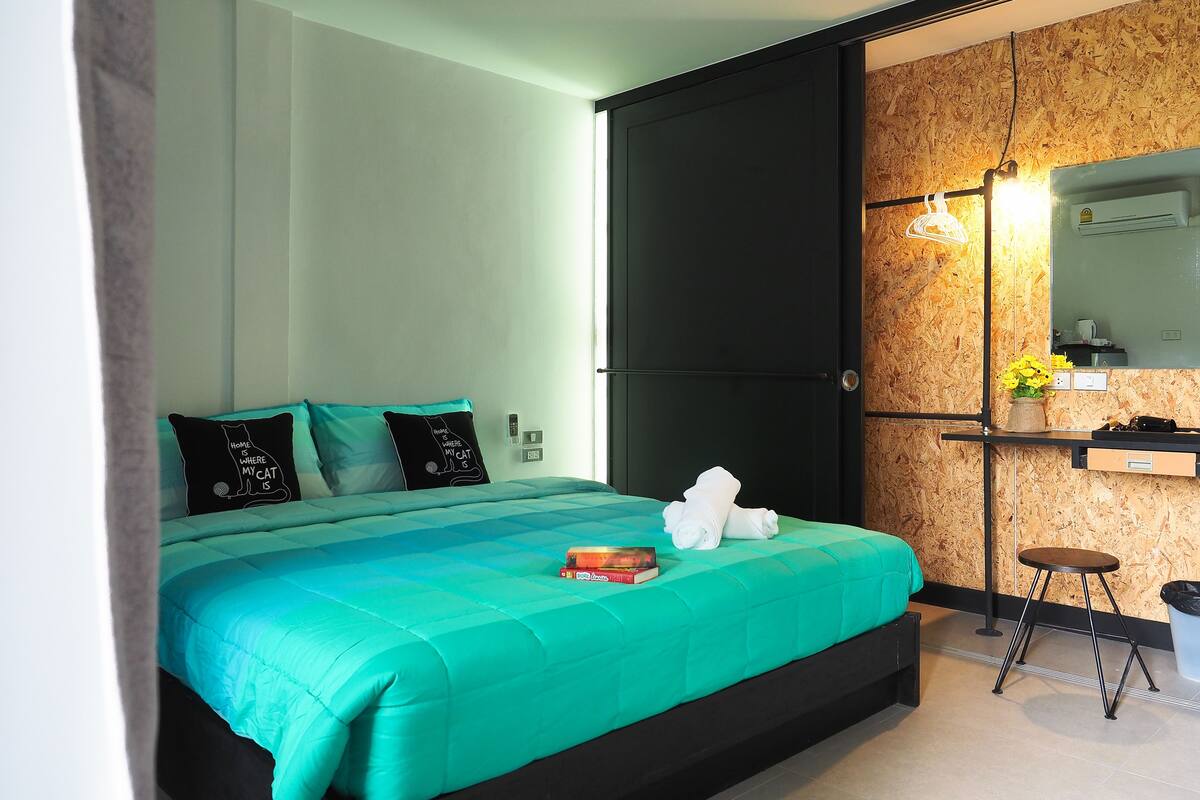 A modern bedroom with a teal bedspread, black and teal pillows, a small bedside table with a book, and a mirror with a wooden backdrop.