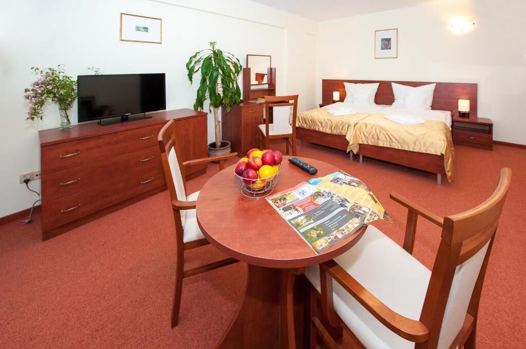 A hotel room with a bed, bedside table, TV, wooden dresser, dining table with chairs, and a fruit bowl.