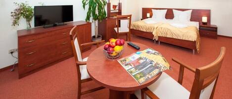 A hotel room with a bed, bedside table, TV, wooden dresser, dining table with chairs, and a fruit bowl.