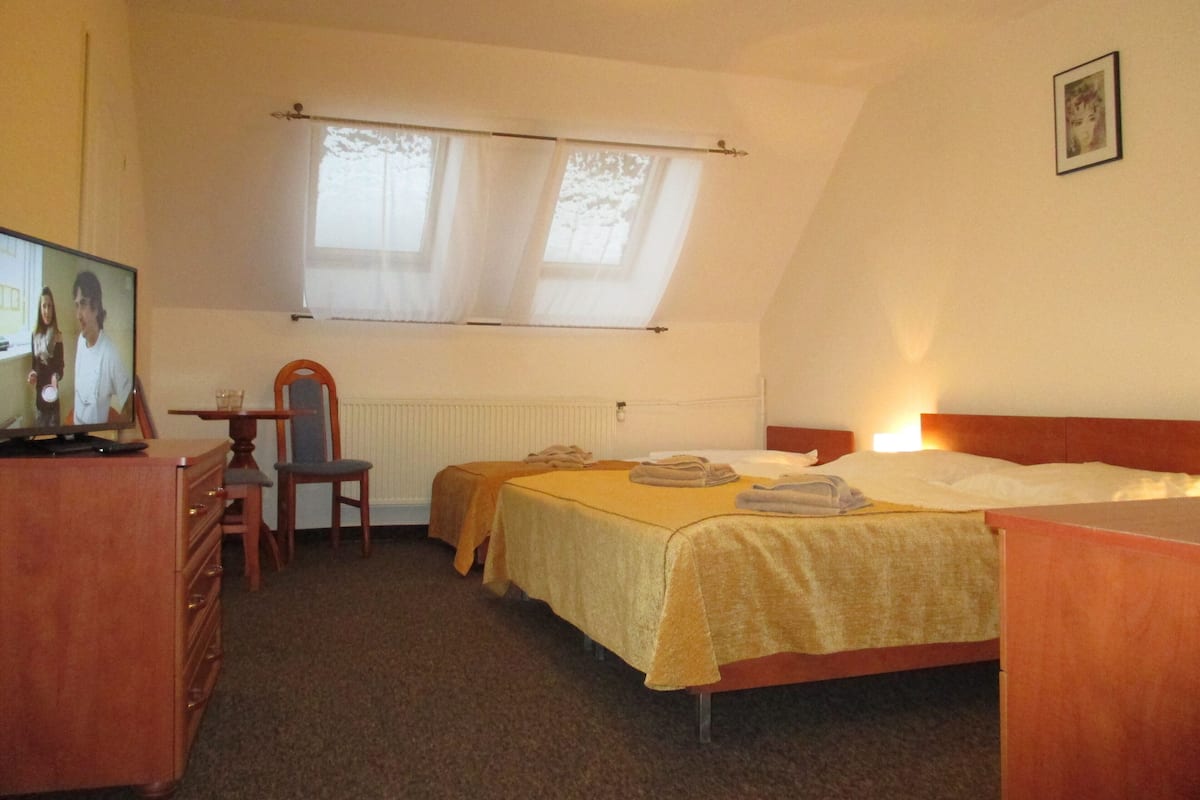 Een slaapkamer met een bed, een bureau met een computer, een stoel en een raam met dunne gordijnen.