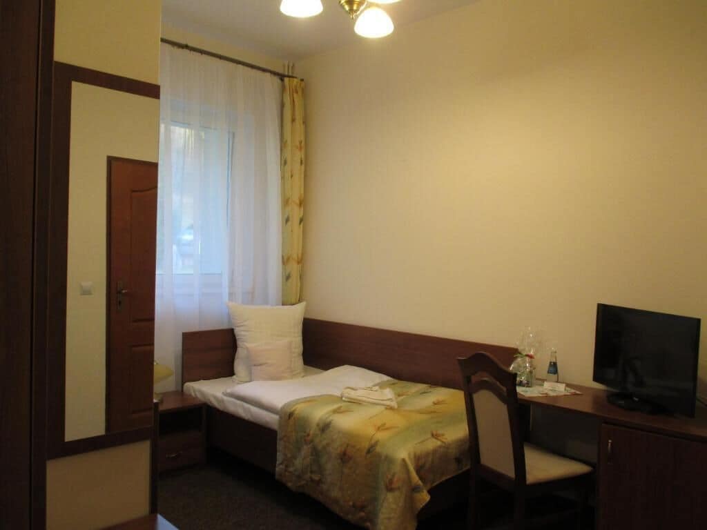 A small, single-bed room with a wooden headboard, a desk with a television, and a window with sheer curtains.