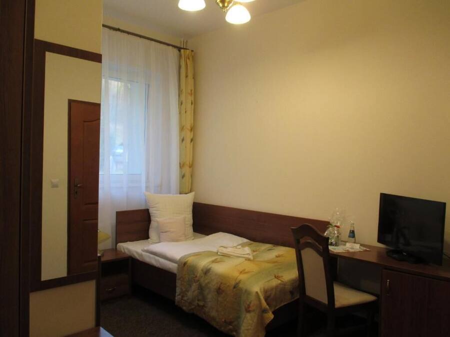 A small, single-bed room with a wooden headboard, a desk with a television, and a window with sheer curtains.