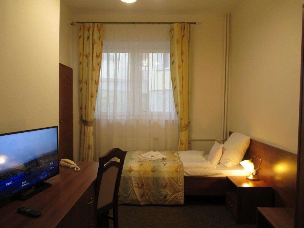 A hotel room with a bed, a desk with a television, and a bedside table with a lamp.