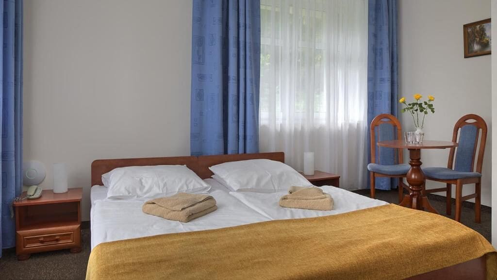 A hotel room with a bed, a nightstand, a chair, and a small table with a vase of flowers.