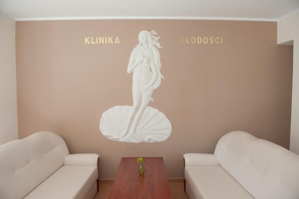 Ein Zimmer mit einer beigen Wand, das eine weiße Skulptur einer Frau zeigt. Auf der Wand sind die Worte "KLINIKA" und "MŁODOSCI" zu sehen. Zwei weiße Sofas und ein Holztisch mit einer Vase in der Mitte ergänzen das Bild.