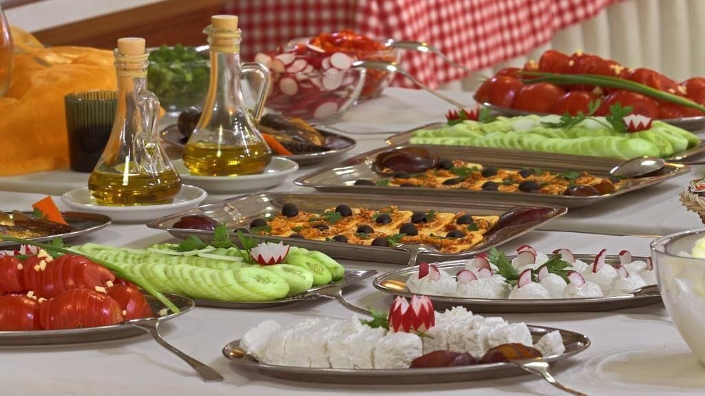 A buffet table with various dishes, including sliced cucumbers, tomatoes, olives, and a variety of cheeses.