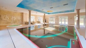 Una amplia piscina interior con un mural en el techo, ventanas grandes y una zona de estar con sillas y mesas.