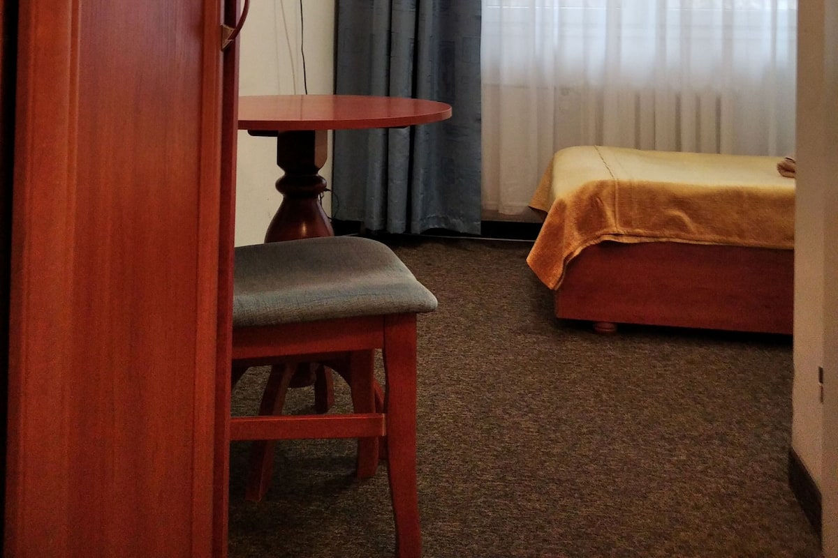 Een kleine kamer met een houten deur, een tafel met een stoel, een bed met een gele deken, een televisie aan de muur en een raam met gordijnen.
