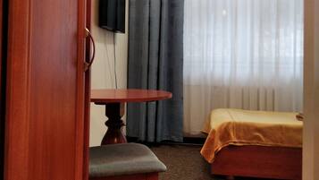 Una habitación pequeña con puerta de madera, una mesa con una silla, una cama con una manta amarilla, un televisor fijado a la pared y una ventana con cortinas.