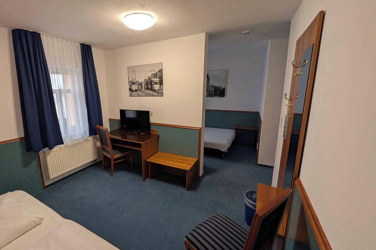 Habitación de hotel con cama, escritorio con computadora, silla, mesita, radiador, ventana con cortinas y puerta con espejo.