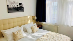 Una camera d'hotel con un letto, un comodino, una lampada, una finestra con tende e un quadro appeso al muro.