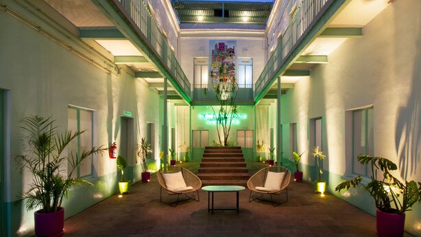 Un vestíbulo de hotel con una escalera central, dos zonas de estar con sillones y plantas en macetas.