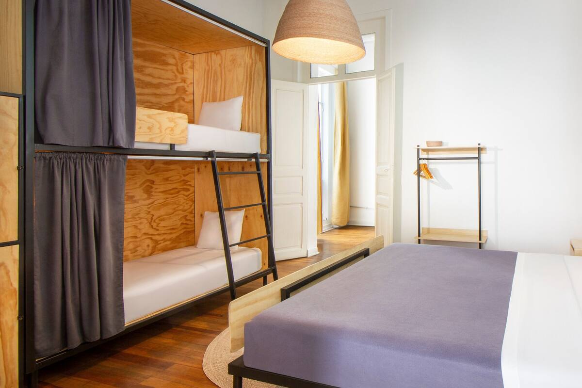 A room with a bunk bed, a single bed, a wooden ladder, a small table, and a hanging light fixture.