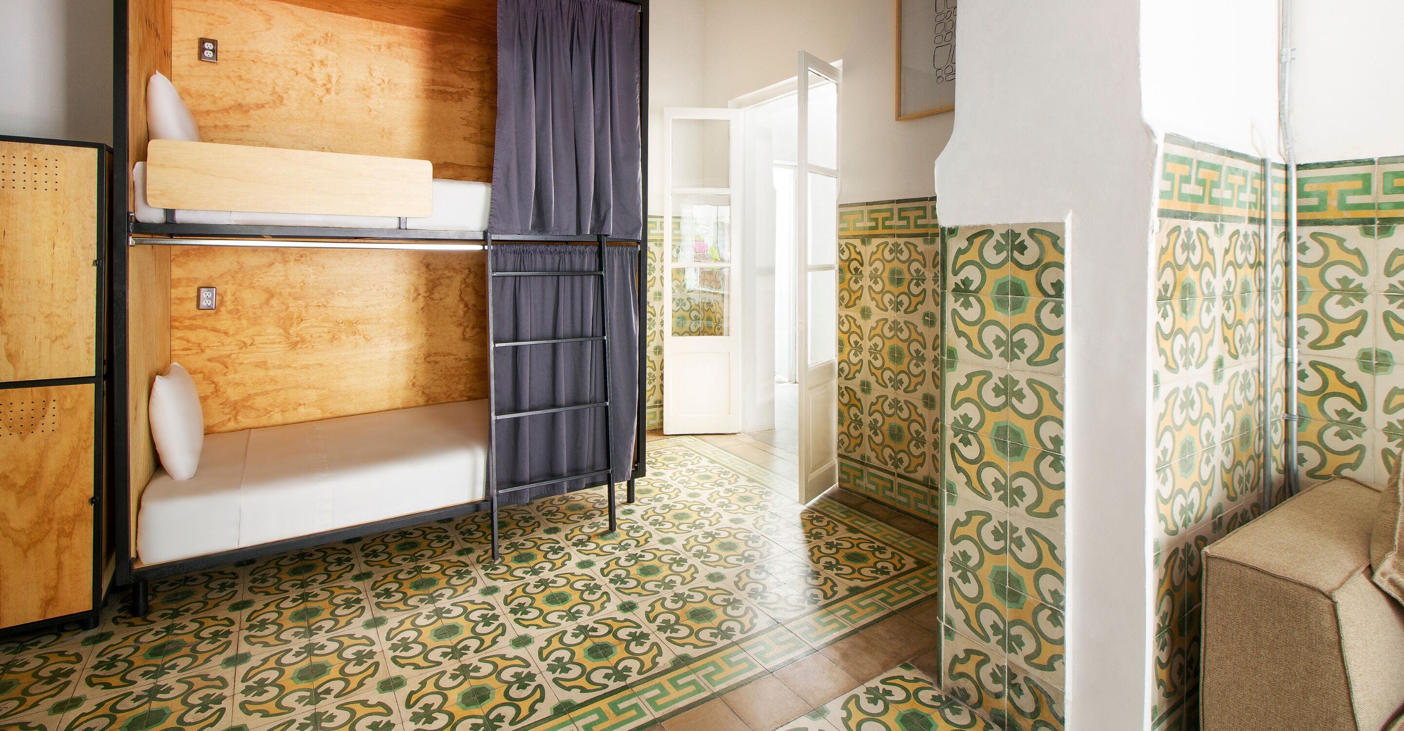 Una habitación con literas, un mueble pequeño, un baño con azulejos estampados y vistas por una puerta abierta.