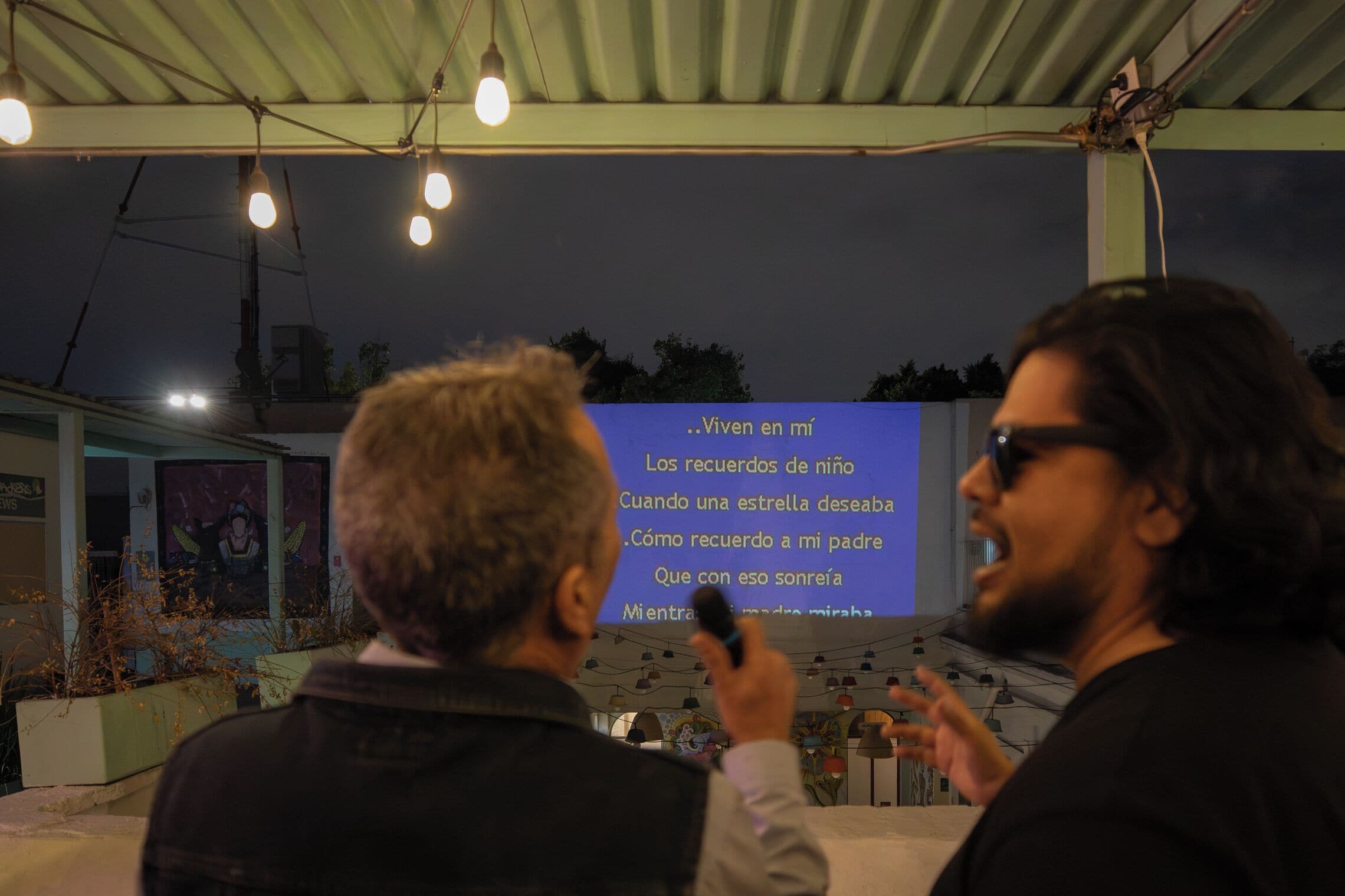 Duas pessoas em ambiente externo com uma tela de projeção exibindo texto.