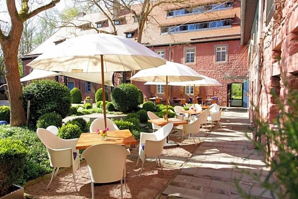 Un área de estar al aire libre con mesas y sillas, rodeada de follaje y un edificio al fondo.