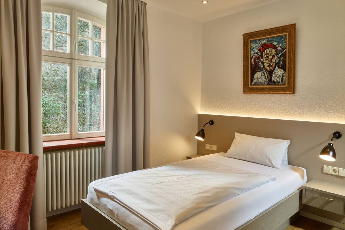 Un lit bien fait, avec du linge de lit blanc, une fenêtre avec des rideaux, un radiateur, une chaise et un tableau accroché au mur.