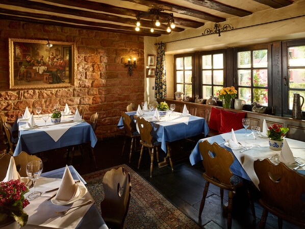 A restaurant with stone walls, wooden beams, and large windows. Tables are set with white tablecloths and napkins. -  Hotel & Restaurant Burgschänke (Kaiserslautern)