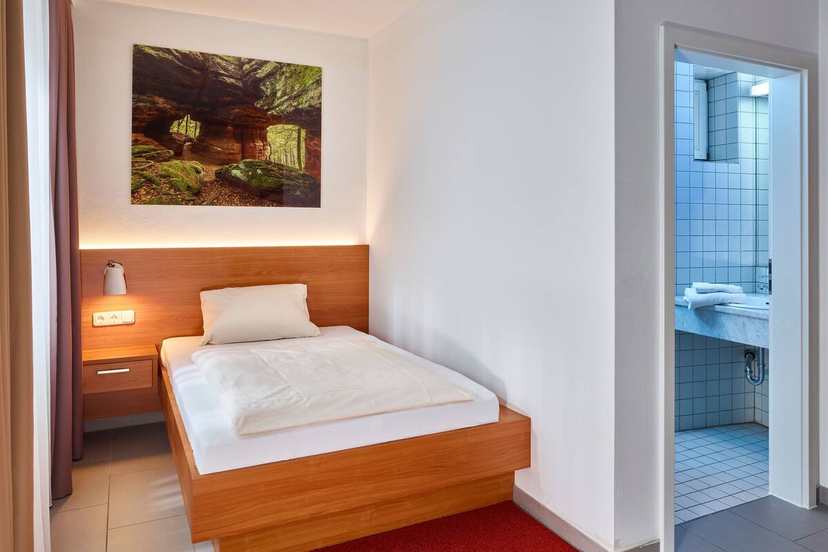 Une chambre d’hôtel avec un lit en bois, une moquette rouge, un tableau représentant une forêt et une salle de bain carrelée de bleu.