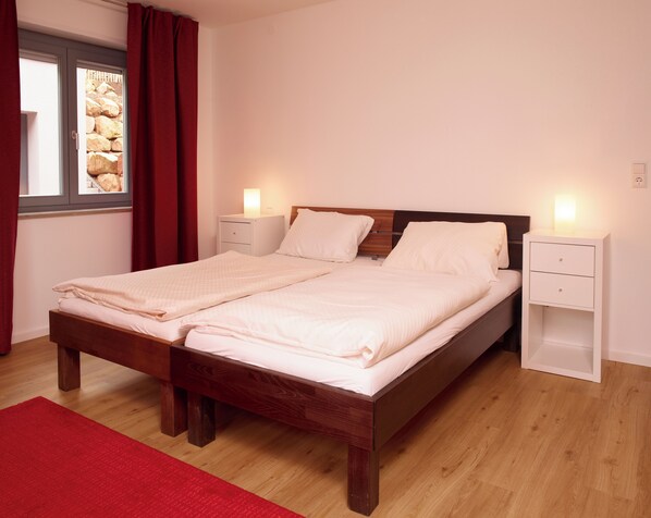 Una cama doble con estructura de madera, dos mesitas de noche con lámparas, una alfombra roja y una ventana con cortinas.