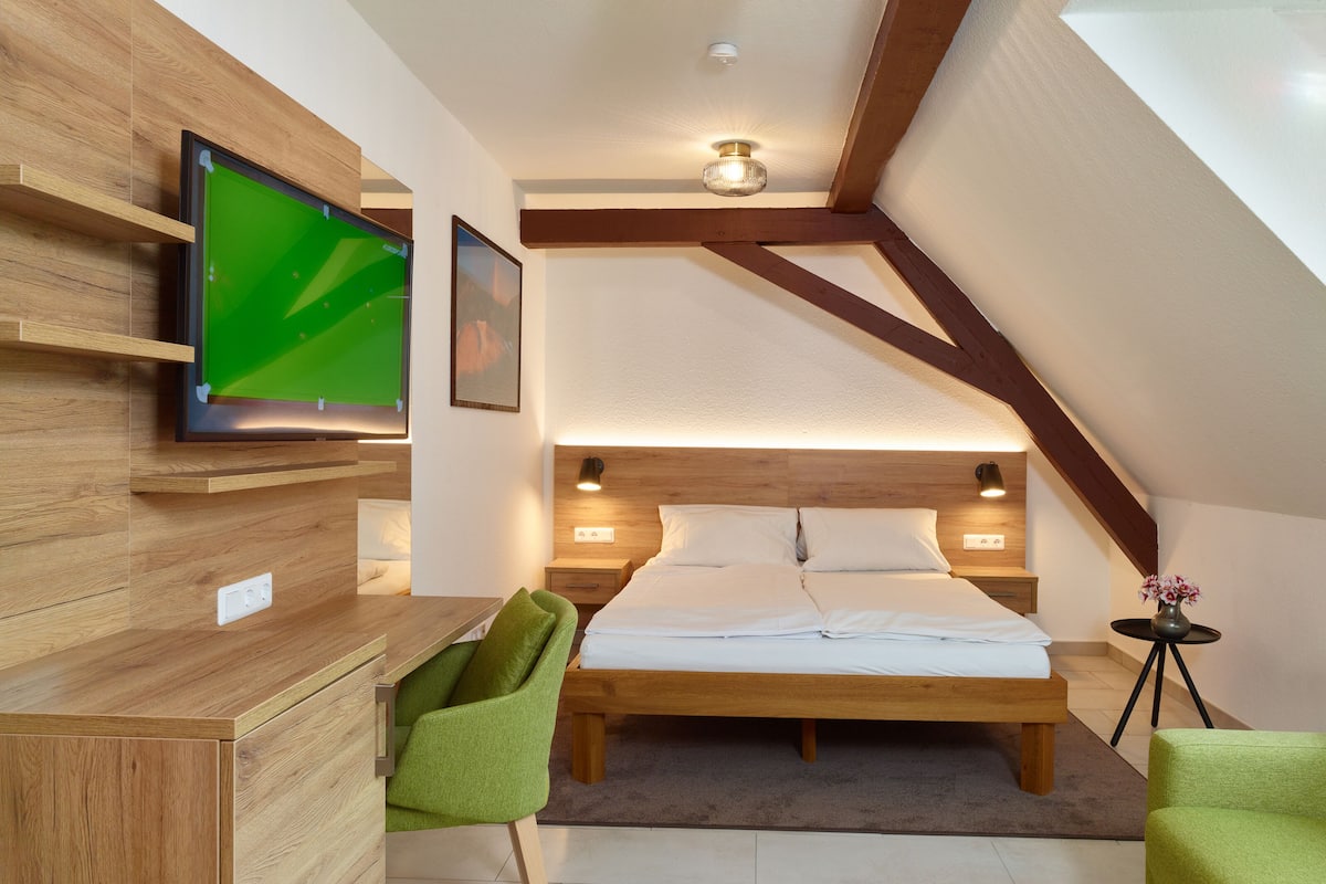 Une chambre moderne avec un bureau en bois, une chaise verte, un lit avec du linge de lit blanc et un téléviseur fixé au mur.