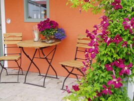 Un área de estar al aire libre con una mesa y sillas, una maceta con flores moradas y una pared con un ventanal.