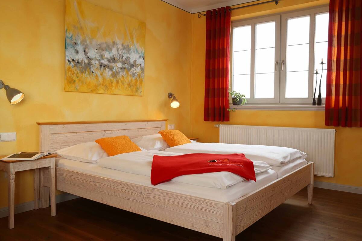 A room with a wooden bed, two bedside lamps, a window with red curtains, and a yellow wall with an abstract painting.