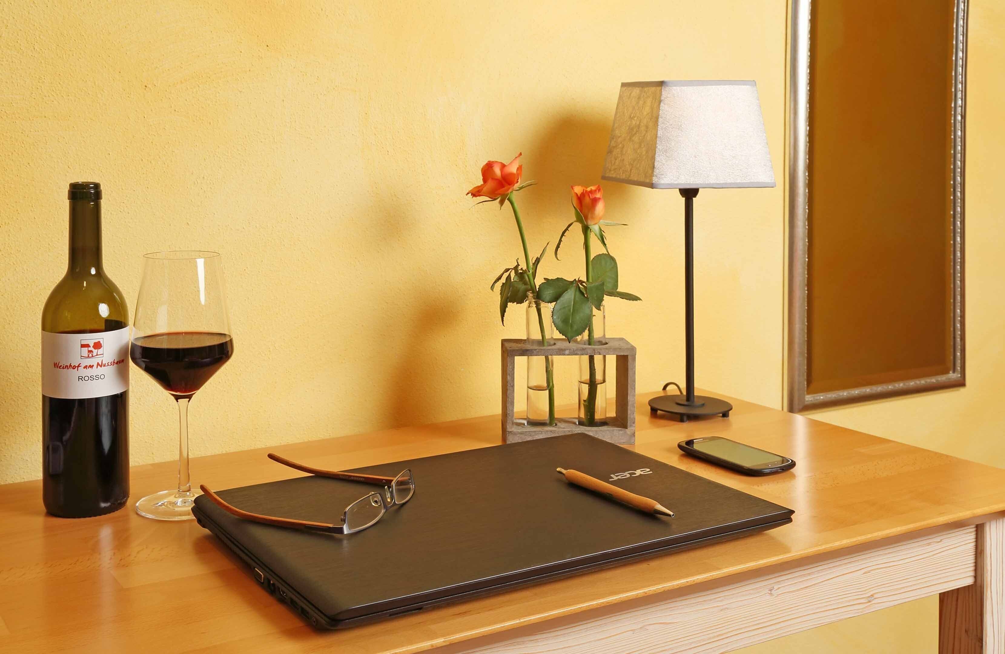 A wooden desk with a laptop, a wine bottle, a glass of red wine, a pair of glasses, a smartphone, a lamp, and two roses in a vase.