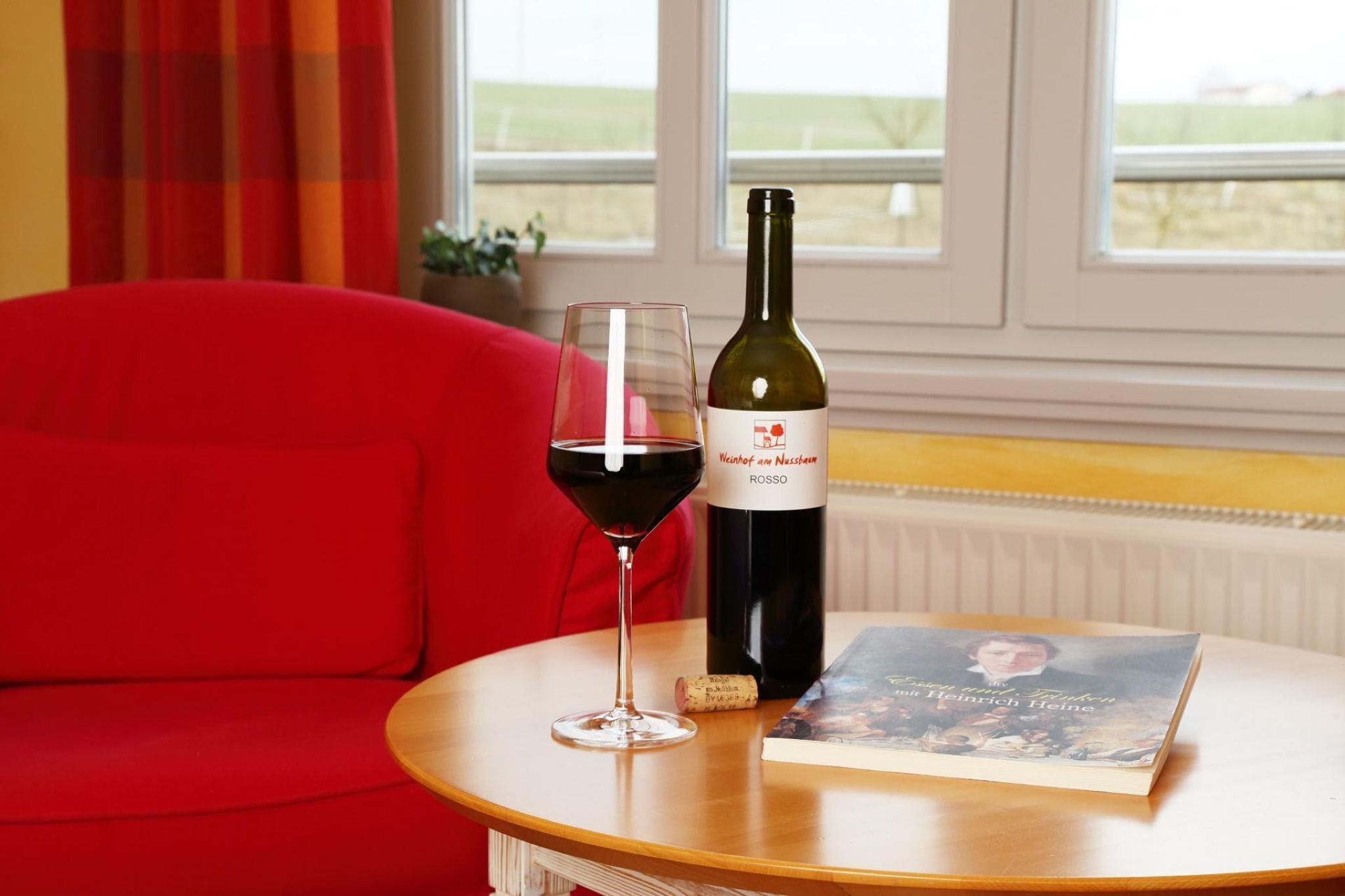 Ein rotes Sofa, eine Weinflasche, ein Weinglas und ein Buch auf einem Tisch neben einem Fenster.