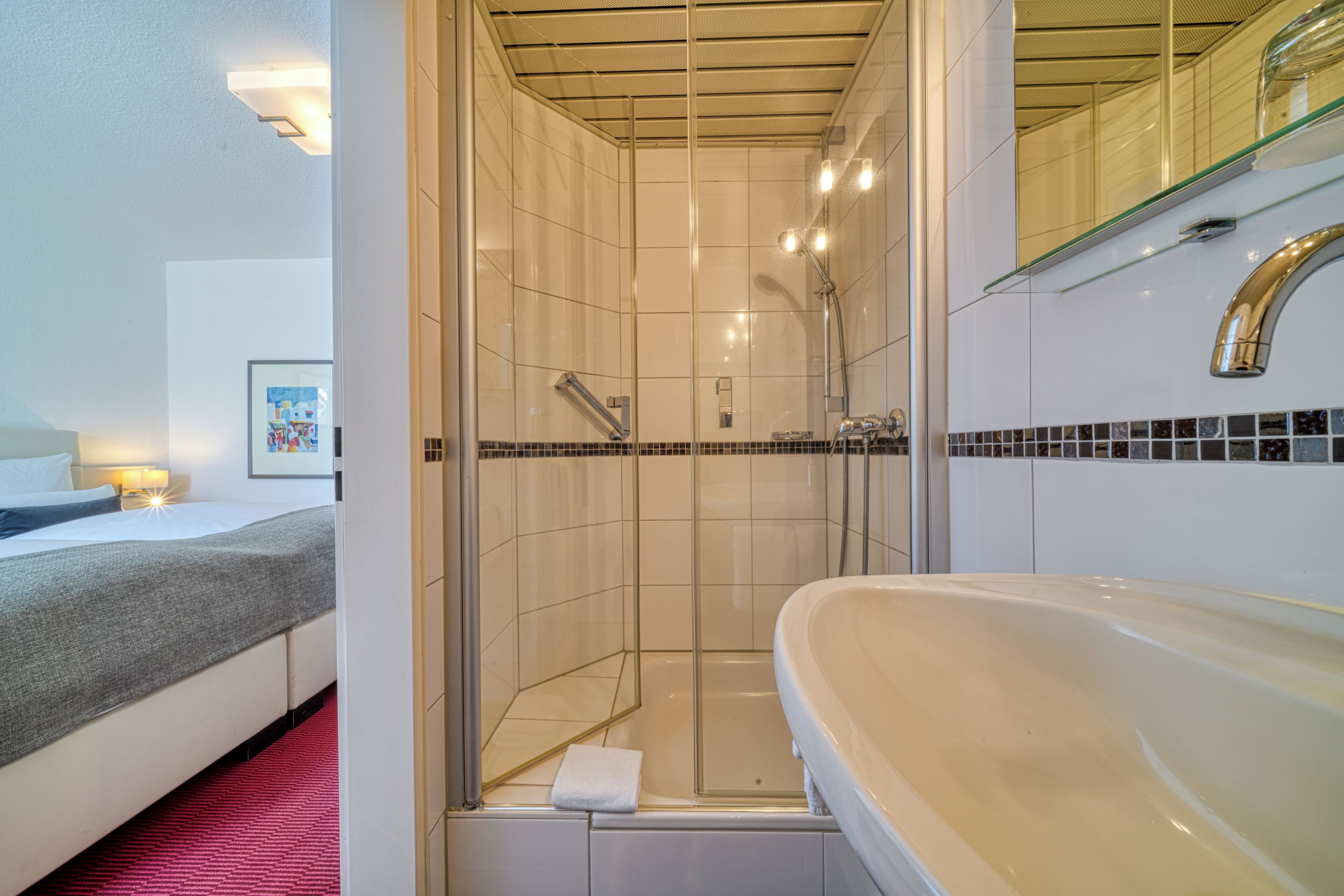 A hotel bathroom with a shower, a sink, and a bed with a red carpet.