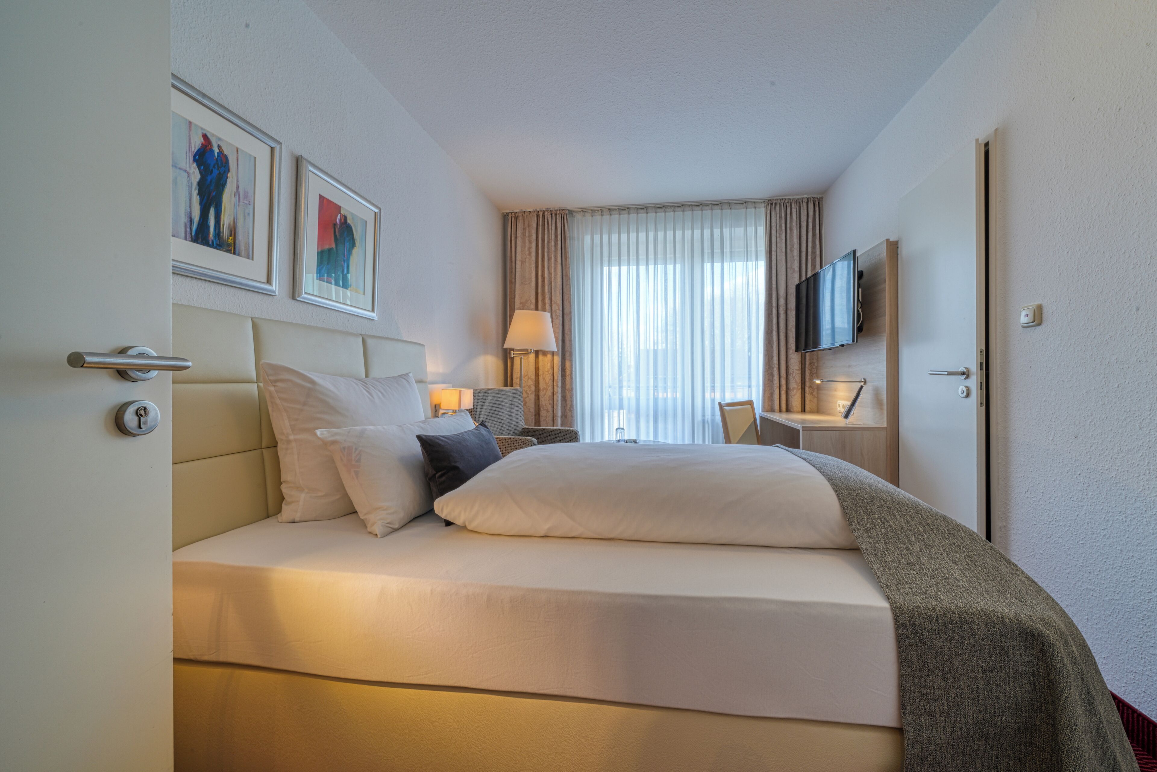 A hotel room with a bed, a television, a desk, and artwork on the wall.
