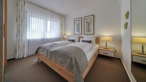 Ein Hotelzimmer mit einem hölzernen Bett, weißen Vorhängen, zwei Wandlampen und zwei gerahmten floralen Bildern.