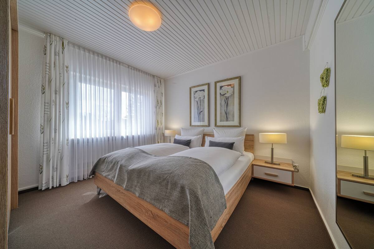 A hotel room with a wooden bed, white curtains, two wall-mounted lamps, and two framed floral paintings.