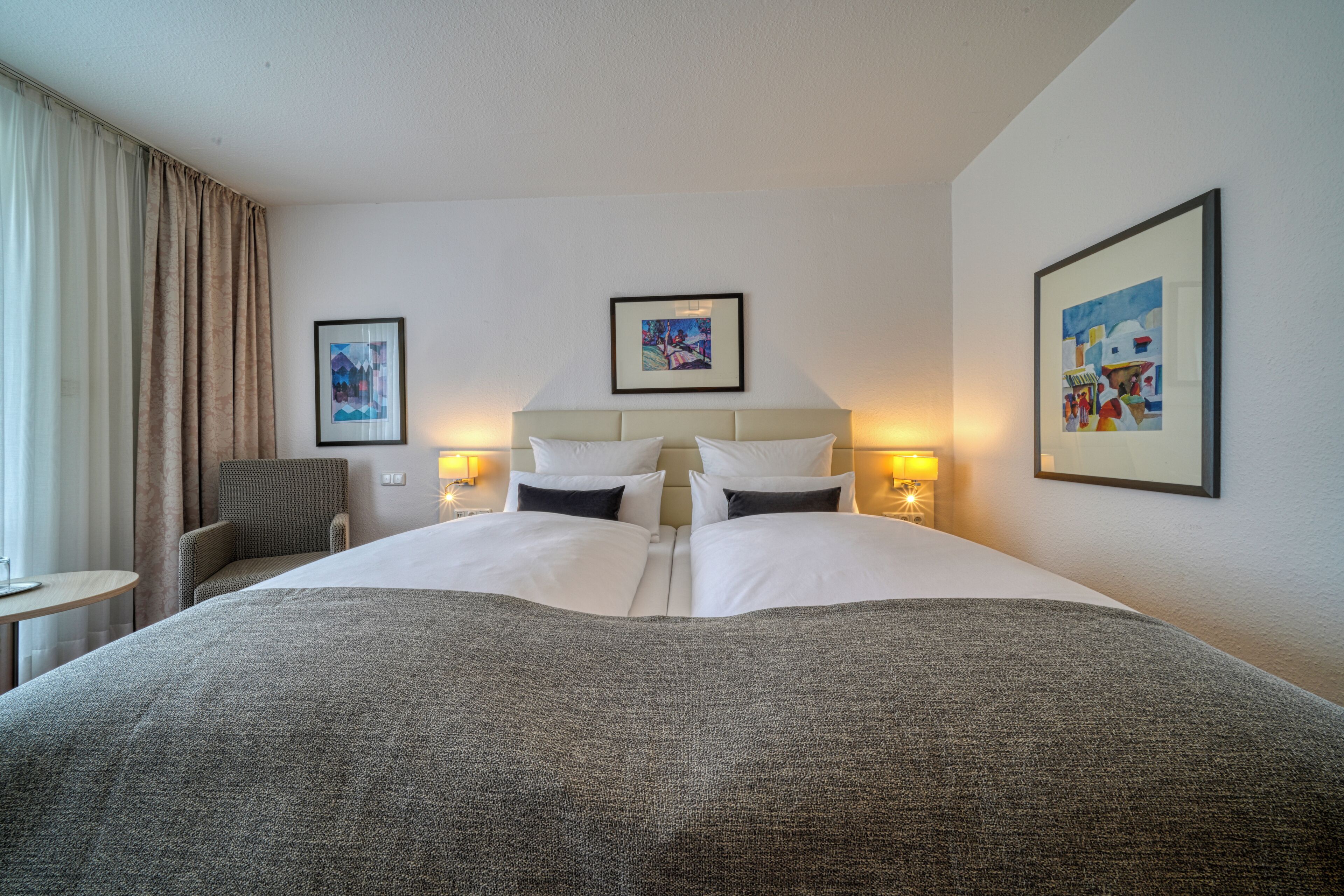 A hotel room with a large bed, two bedside tables with lamps, a chair, and two framed pictures on the wall.