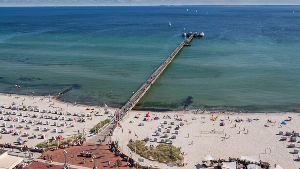 Ein Strand mit einer langen Steg, der sich ins Meer erstreckt, umgeben von Badegästen und Sonnenschirmen.
