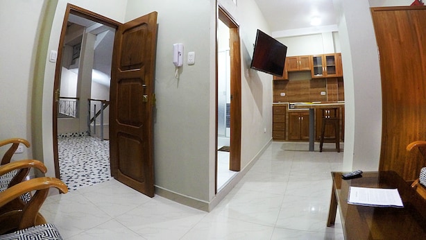 A room with a wooden door, a flat-screen TV mounted on the wall, a kitchen area with wooden cabinets, and a dining table with chairs.